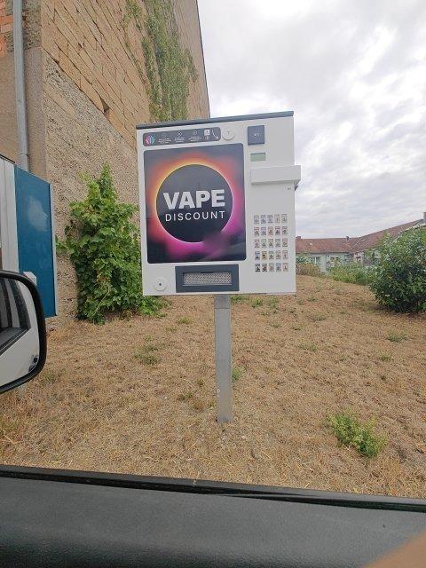 Freistehender weißer Vape-Automat auf einem Pfosten am Ortsrand, großes Logo auf der Front, rechts daneben ein Feld mit kleinen Produktbildern zur Auswahl