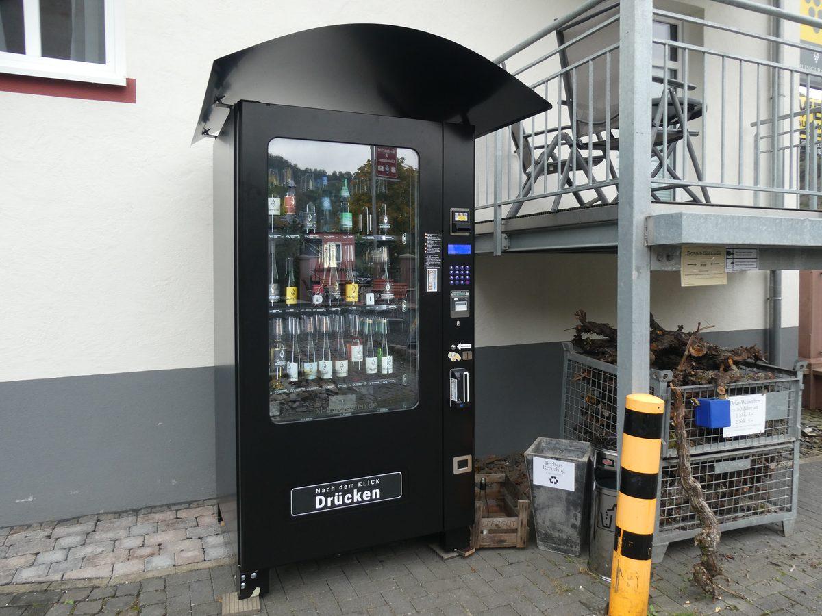 Schwarzer Weinautomat mit Glasfront und gebogenem Vordach an einer hellen Hauswand, hinter dem Glas Weinflaschen in drei Reihen, rechts ein Bedienfeld mit Display, Tastatur und Kartenschlitz, daneben eine Außentreppe und ein Gitterkorb mit Rebholz