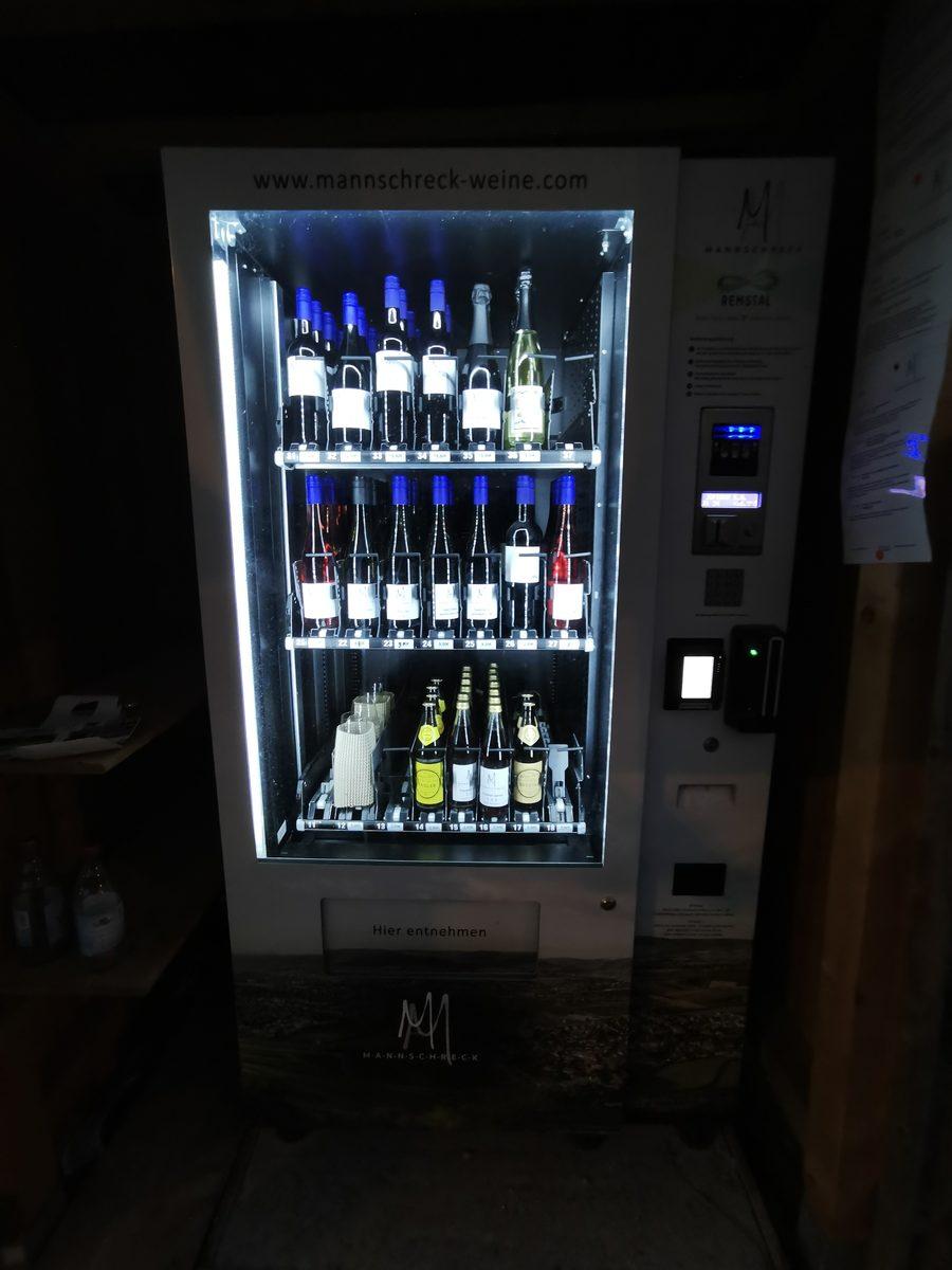 Beleuchteter Weinautomat bei Nacht mit drei Fachreihen voller Wein- und Sektflaschen, oben die Webadresse des Weinguts Mannschreck, rechts ein Bedienfeld mit Display, Kartenterminal und einem zweiten Lesegerät, unten das Ausgabefach mit der Aufschrift Hier entnehmen
