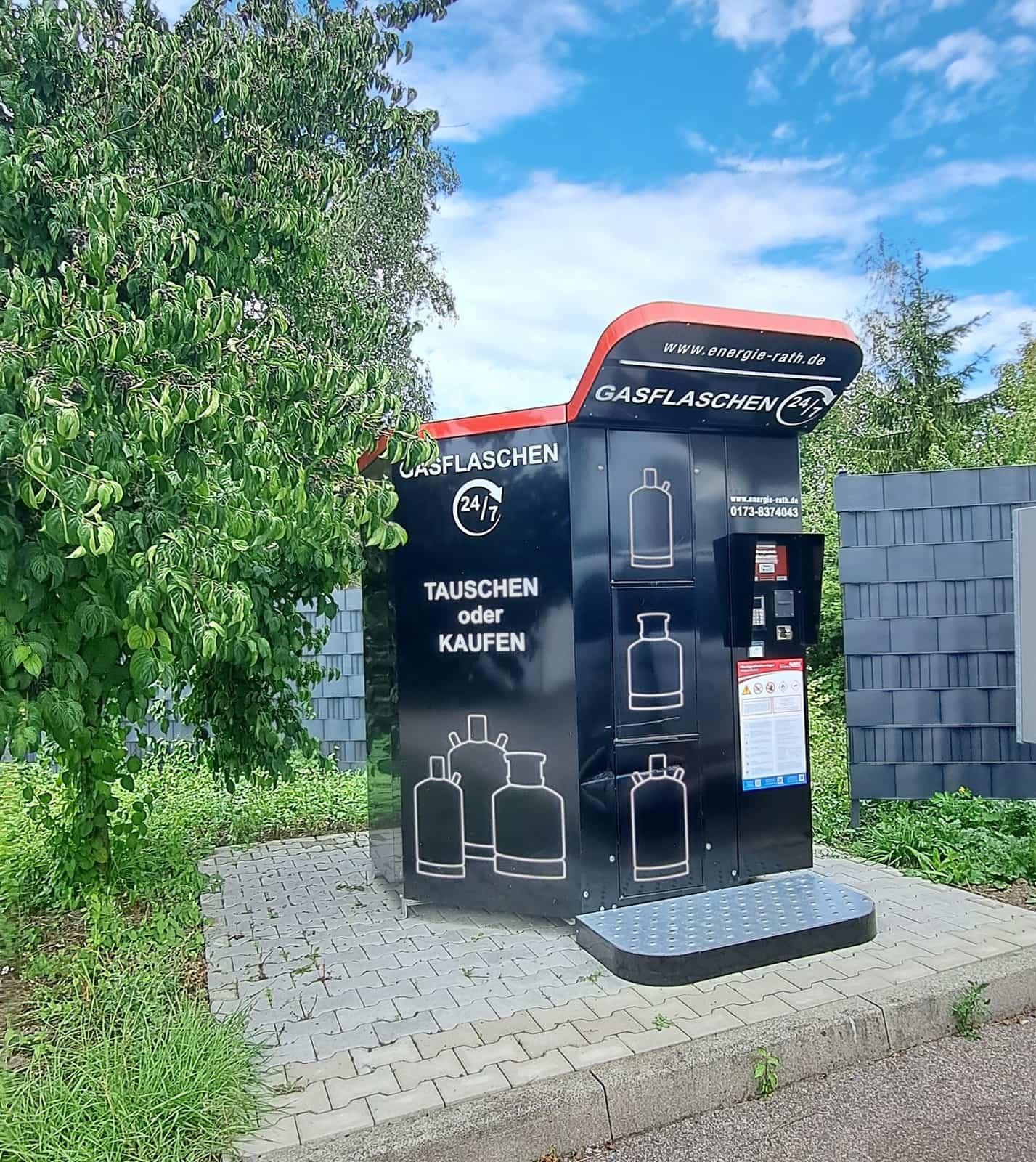 Schwarzer Gasflaschenautomat mit rotem Dach am Straßenrand, dahinter Bäume und Hecke