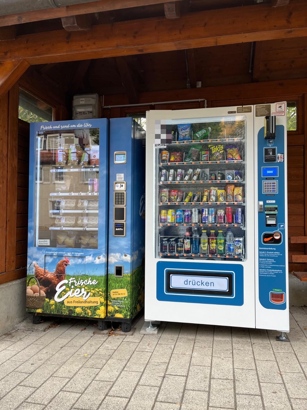 Zwei Verkaufsautomaten unter einem Holzvordach, links ein blau folierter Eierautomat mit Huhn und der Aufschrift Frische Eier aus Freilandhaltung, rechts ein Snackautomat mit Getränken und Süßwaren