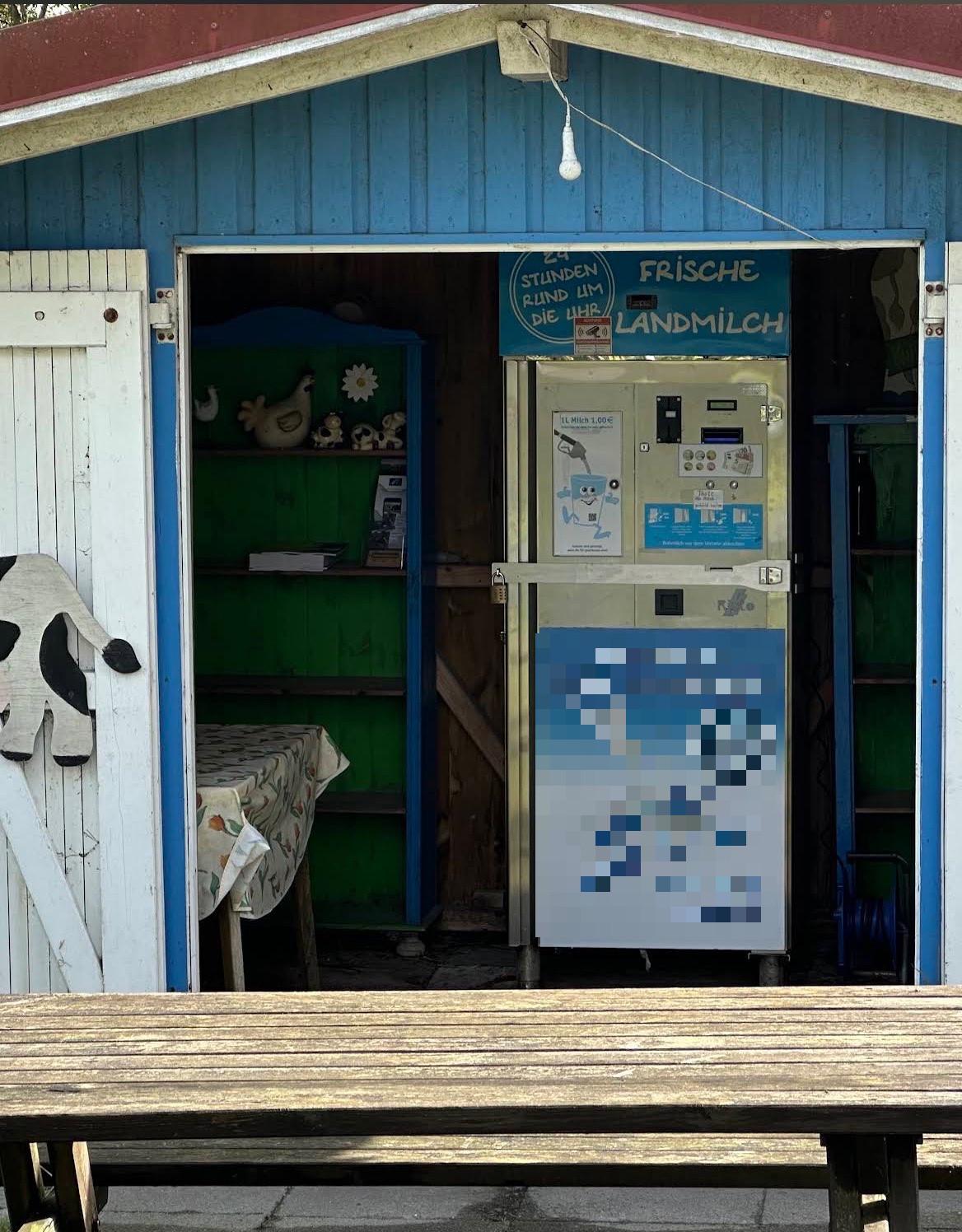 Blaue Holzhütte mit offenem Durchgang, darin ein Milchautomat, davor eine Holzbank