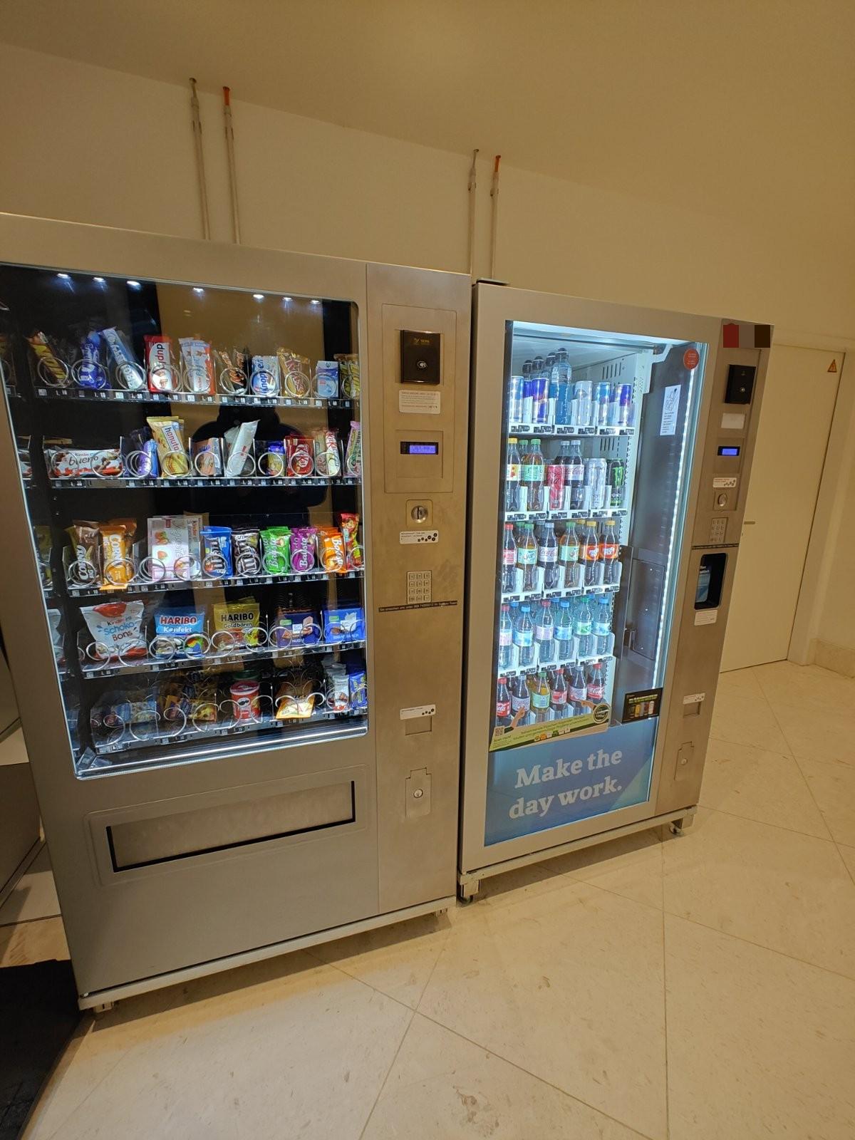 Zwei Verkaufsautomaten in einem Innenraum, links ein Snackautomat mit Bedienfeld aus Kartenleser, Display und Tastatur, rechts ein Getränkeautomat mit beleuchteten Flaschenreihen