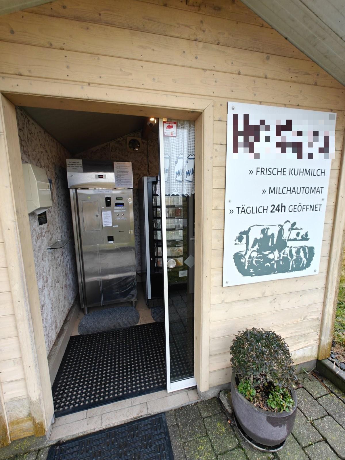 Eingang einer Holzhütte mit Hinweisschild auf frische Kuhmilch und Milchautomat, täglich 24 Stunden geöffnet
